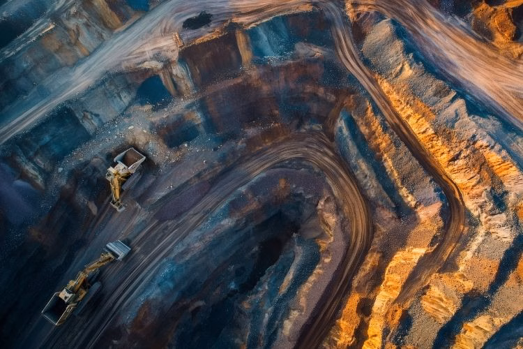 Aerial view of an open-pit mining site with winding roads, excavation equipment, and exposed earth in blue and copper tones.